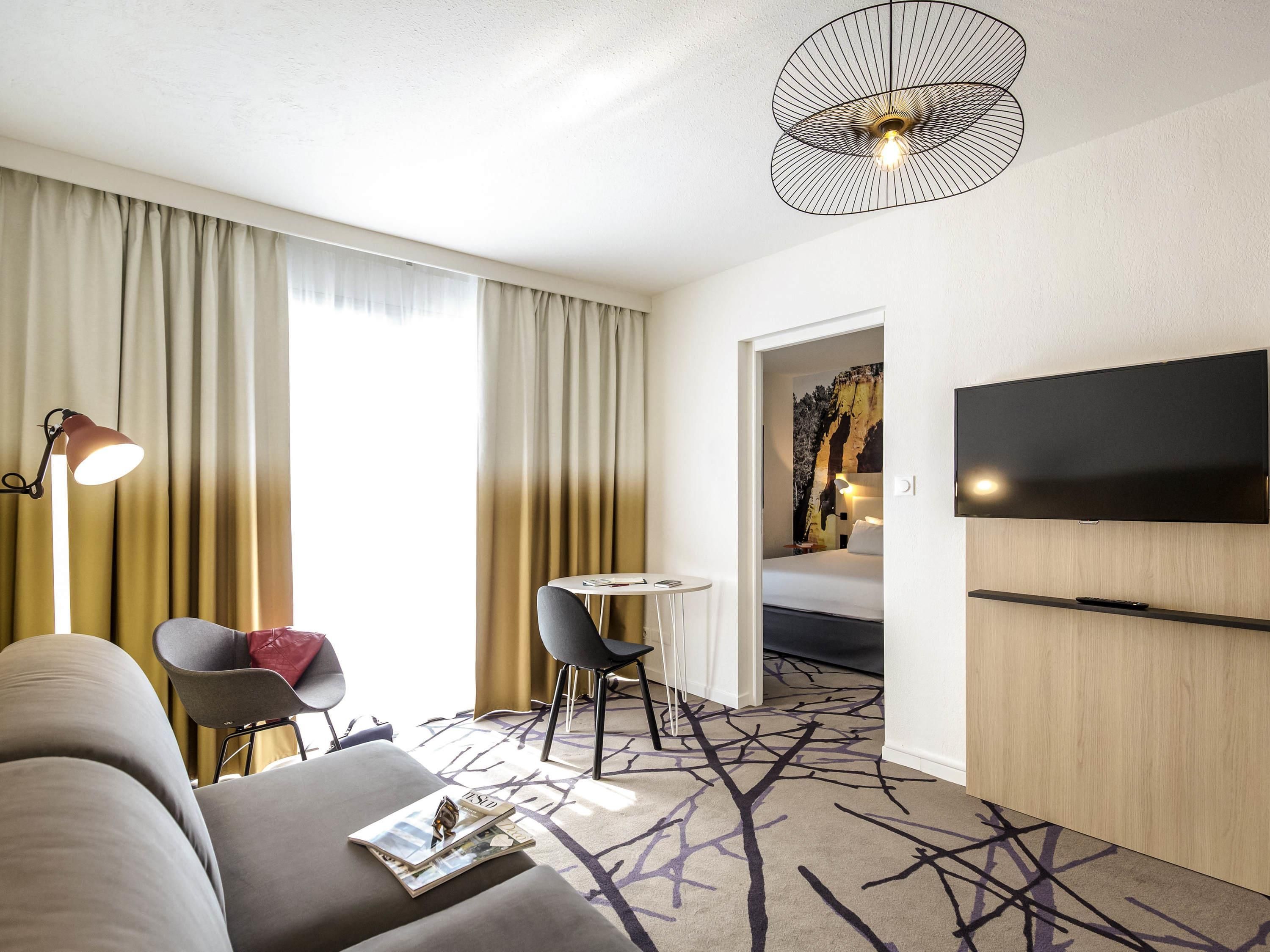 A hotel room with a sofa, a small table, a chair, a lamp, a TV, and a patterned carpet.