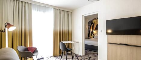 A hotel room with a sofa, a small table, a chair, a lamp, a TV, and a patterned carpet.