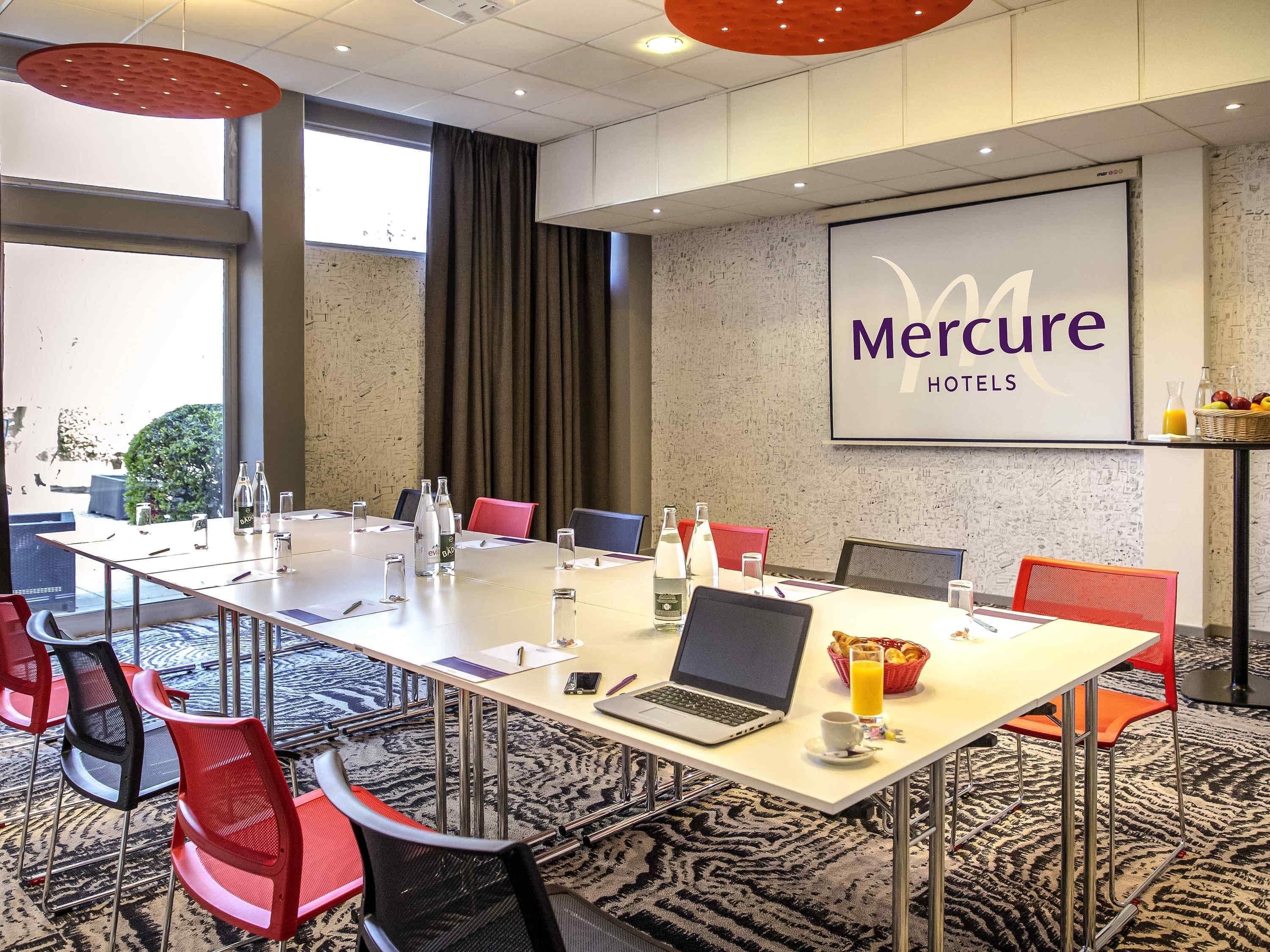 A conference room with a long table, chairs, a laptop, and a projector screen displaying 'Mercure Hotels'.