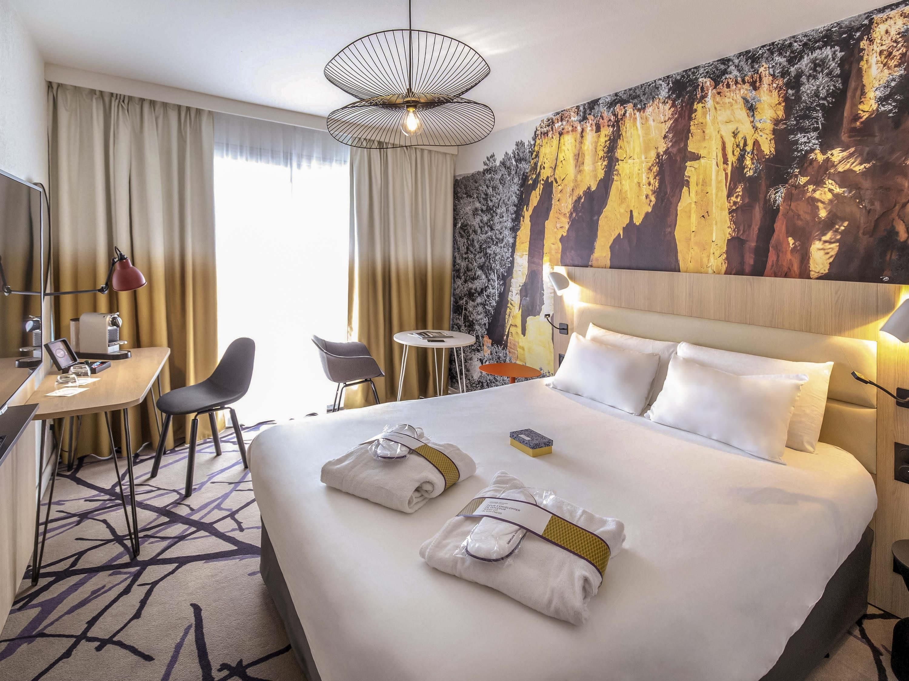 A hotel room with a large bed, a desk with a chair, a small round table, and a wall with a forest mural.