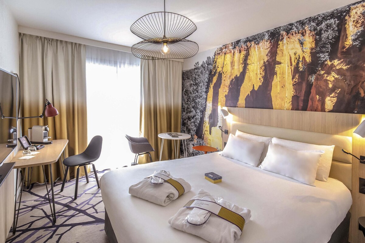 Habitación de hotel con una cama grande, un escritorio con una silla, una mesita redonda y una pared decorada con un mural de un bosque.