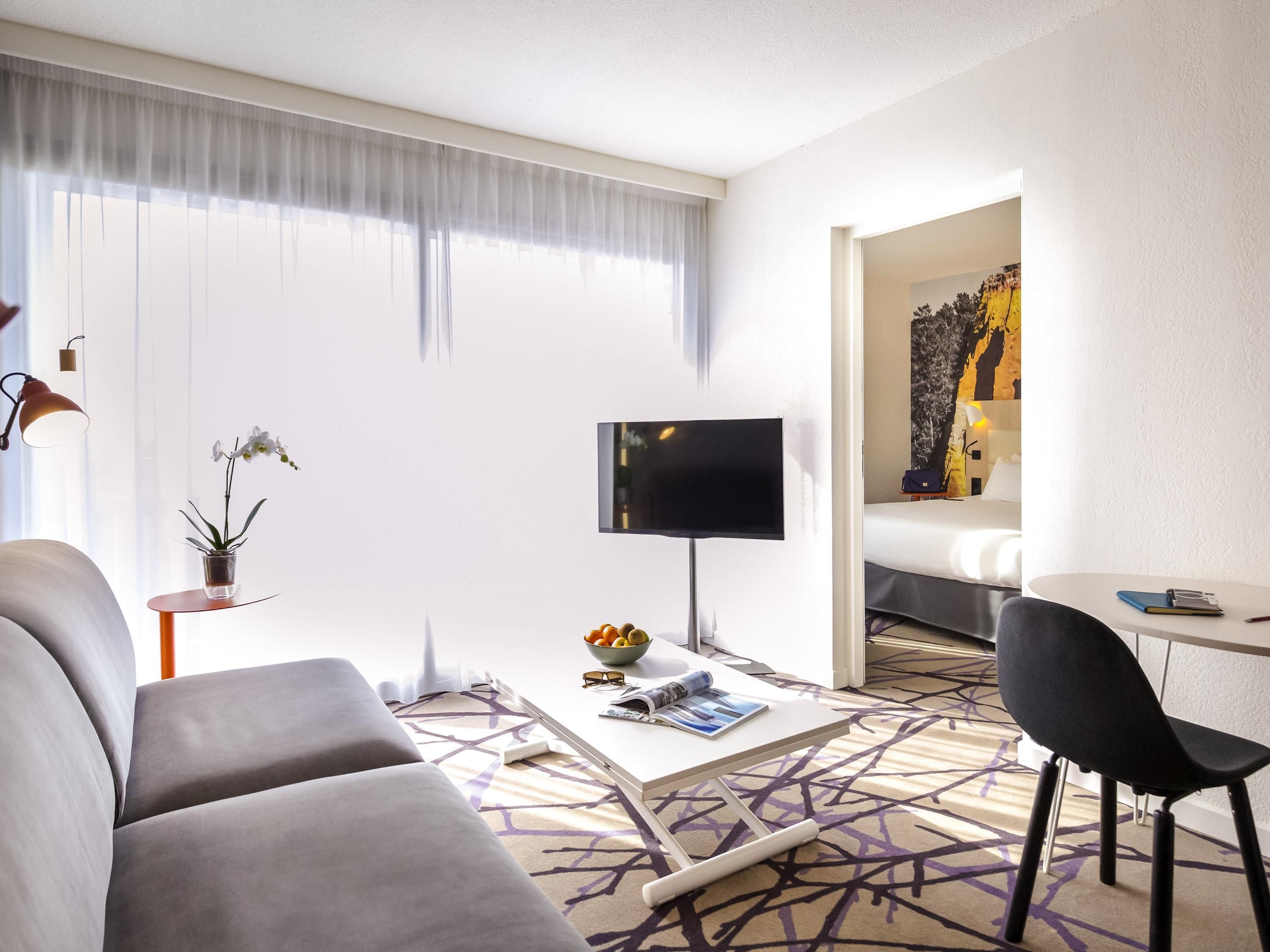 A modern hotel room with a sofa, a coffee table, a TV, and a dining area.