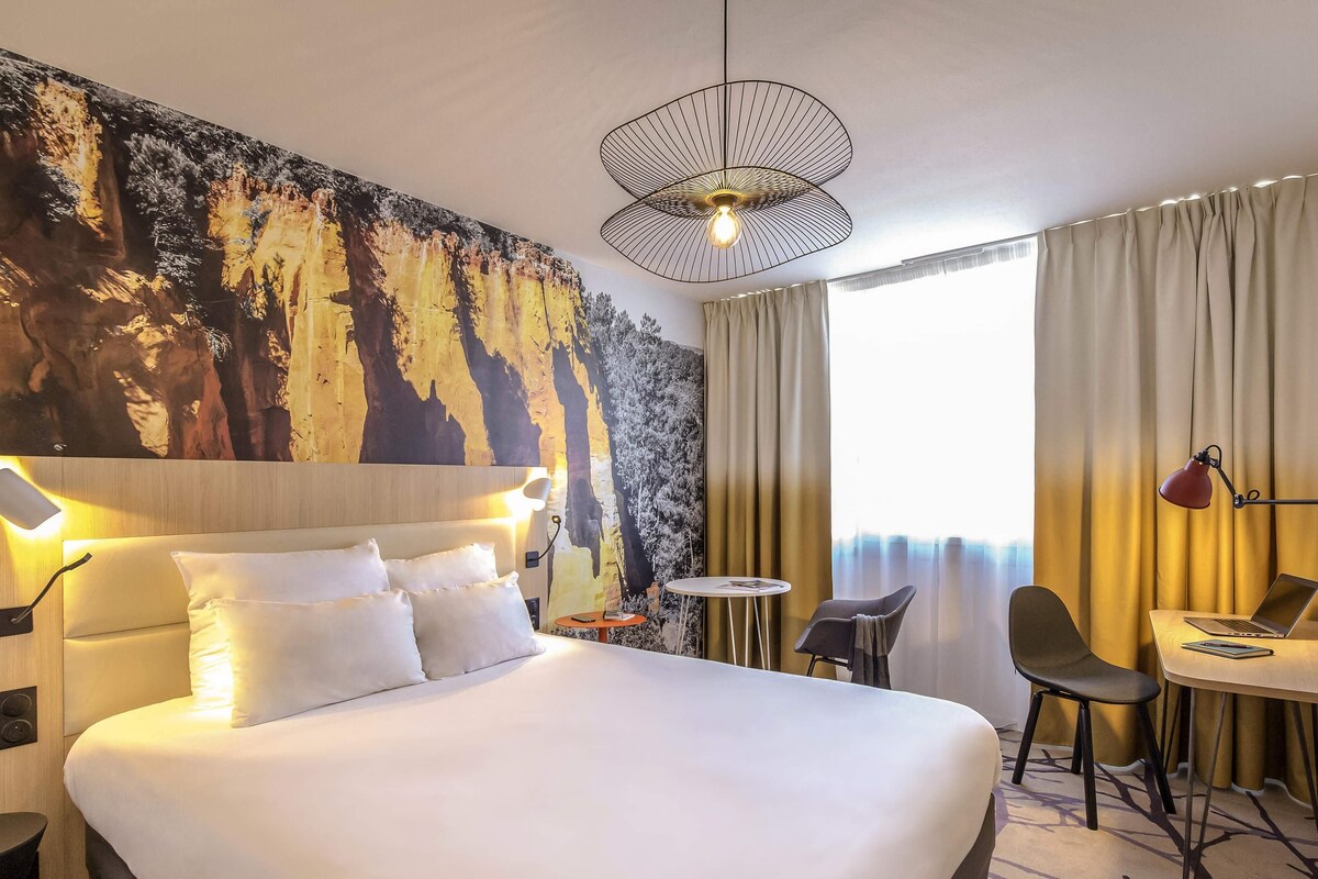 Habitación de hotel con una cama grande, un escritorio con una computadora portátil, una silla, una mesa redonda y un mural original en la pared.