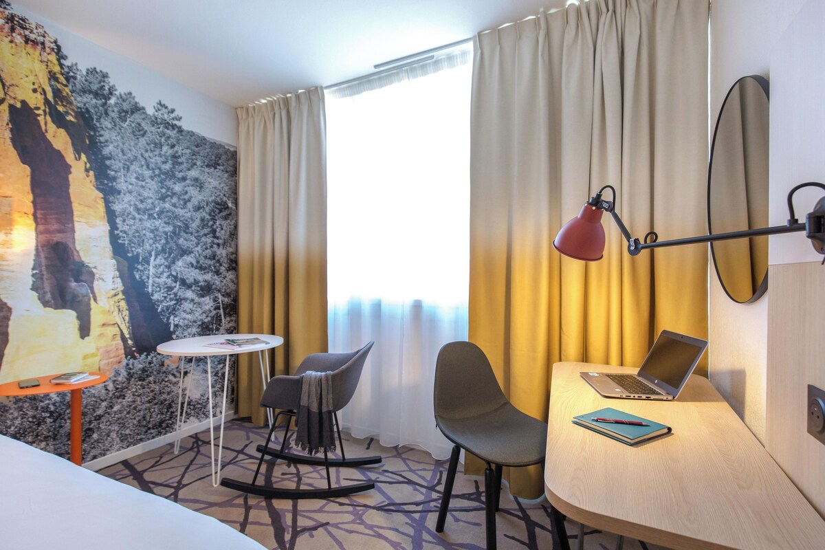 A hotel room with a desk, two chairs, a laptop, a lamp, and a large painting on the wall.