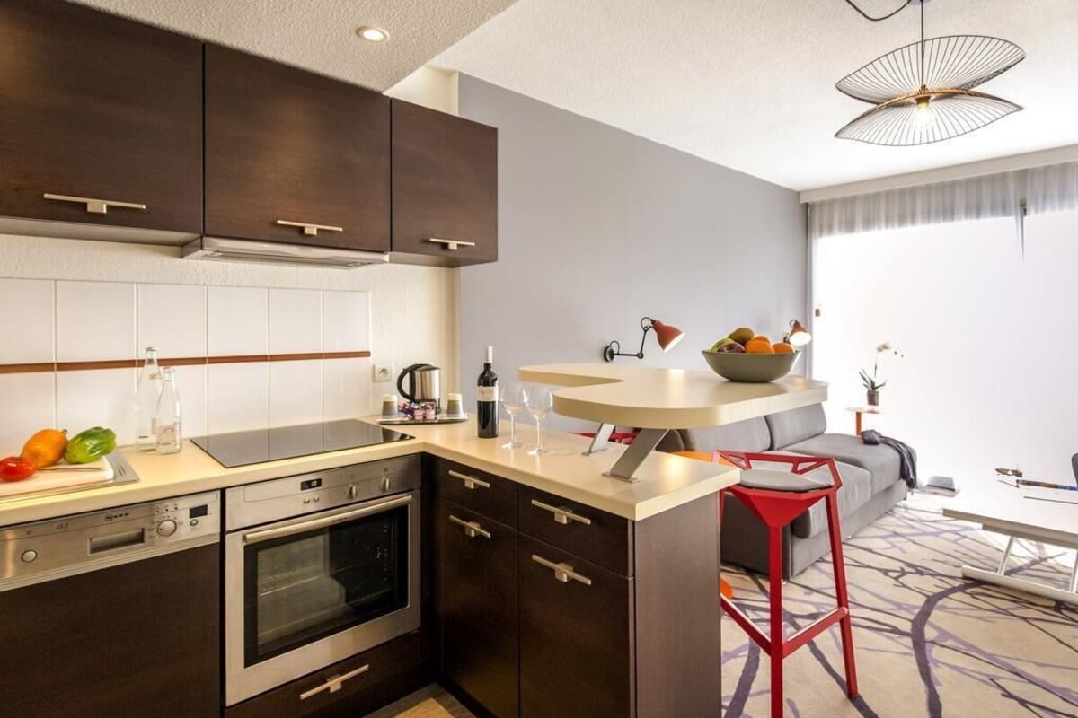 A modern kitchen with dark cabinetry, a built-in oven, a countertop with a wine bottle, glasses, and a bowl of fruit.