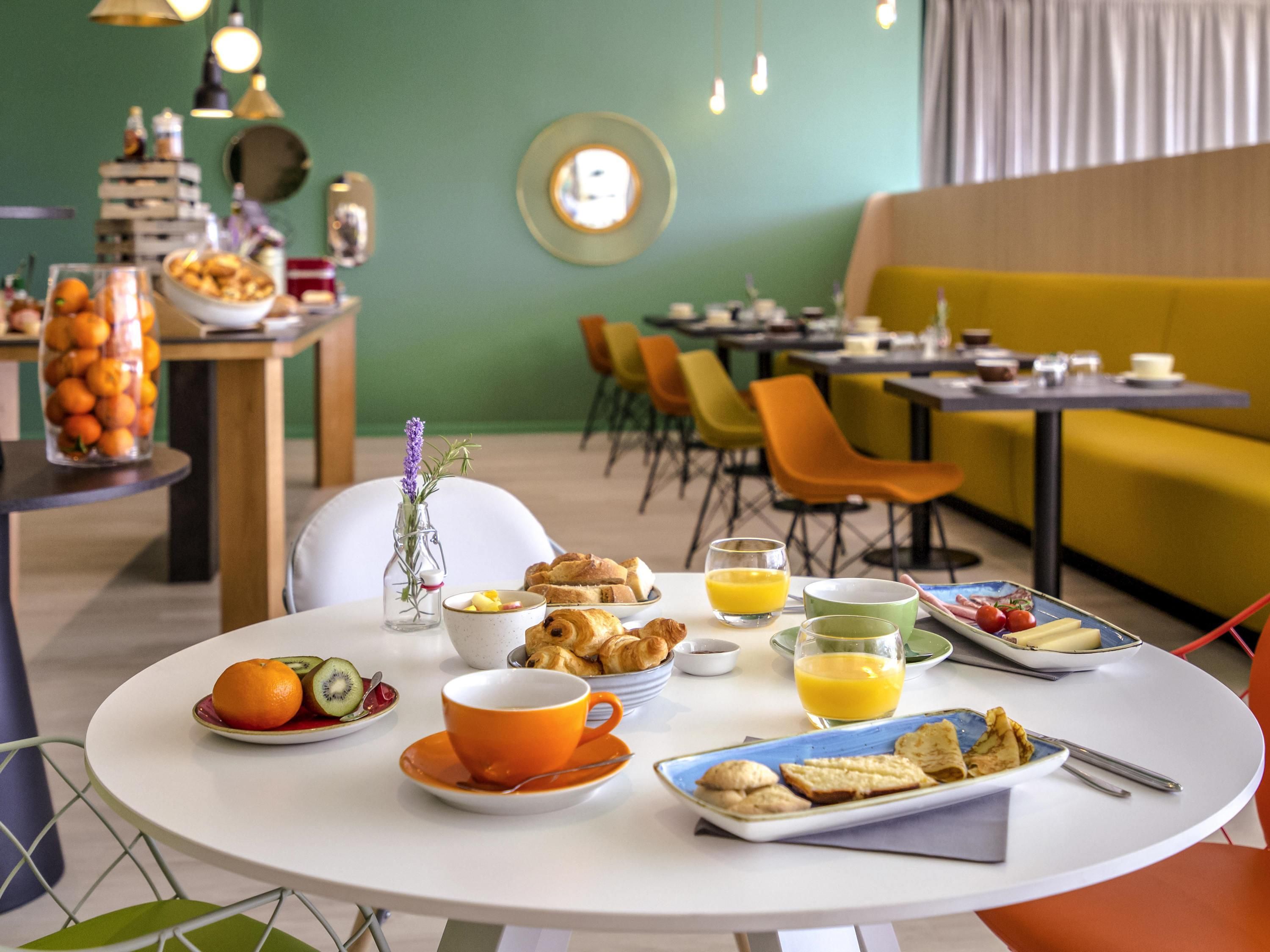 Une table mise pour le petit-déjeuner, avec une nappe pour deux personnes, comprenant des assiettes de fruits, des viennoiseries et un verre de jus d'orange.