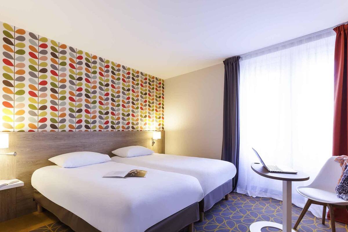 A hotel room with two beds, a desk with a laptop, a chair, and a patterned wall.
