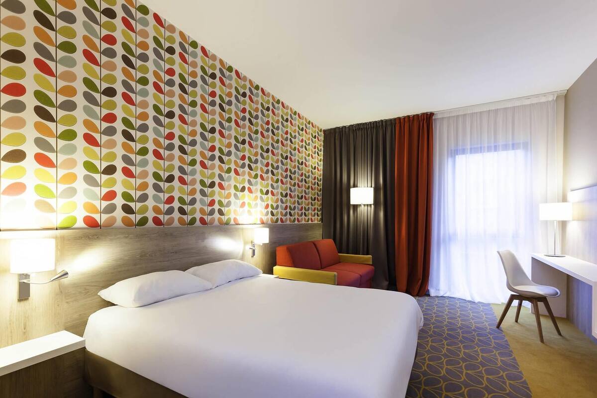 A hotel room with a large bed, a seating area with a chair, a desk, and a wall with a colorful leaf pattern.
