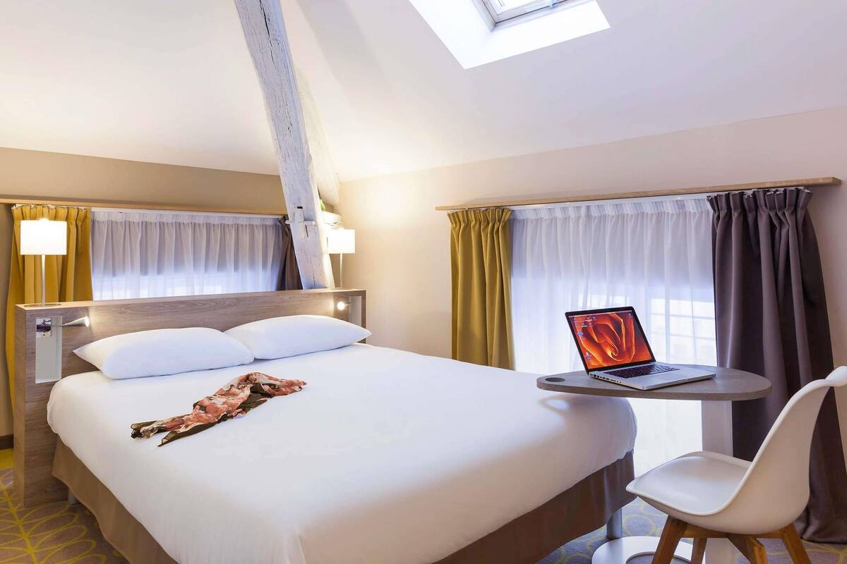 A hotel room with a bed, a desk with a laptop, a chair, and a skylight.