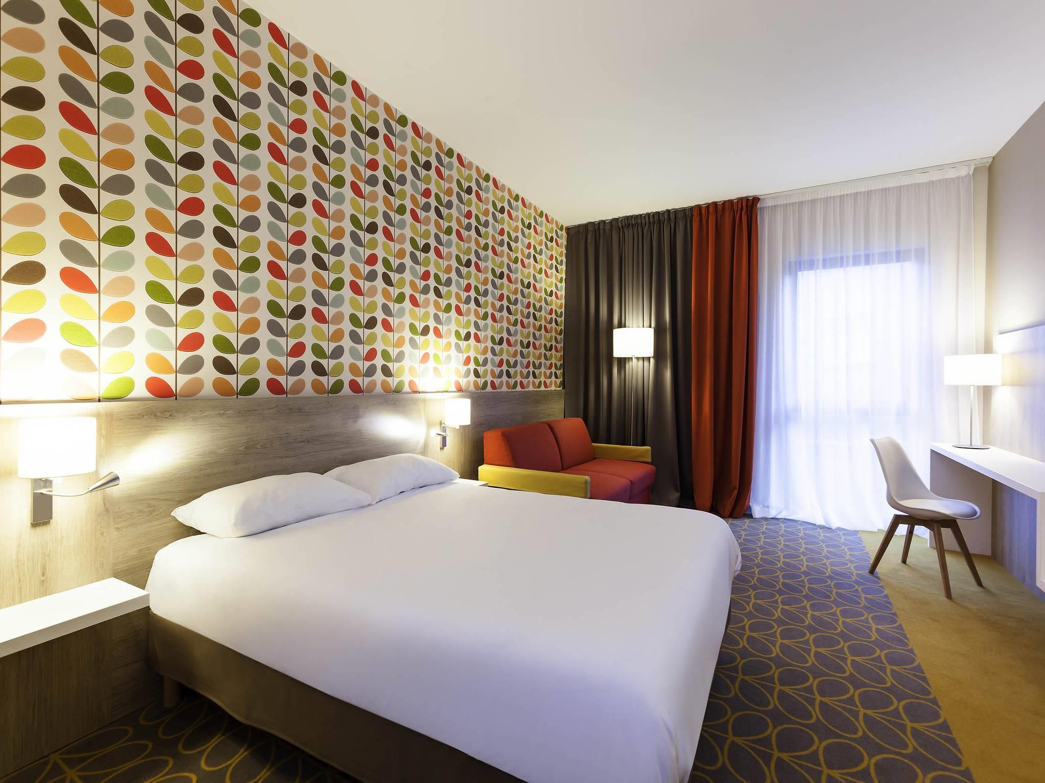 Una habitación de hotel con una cama grande, una zona de estar con una silla, un escritorio y una pared con un diseño de hojas coloridas.