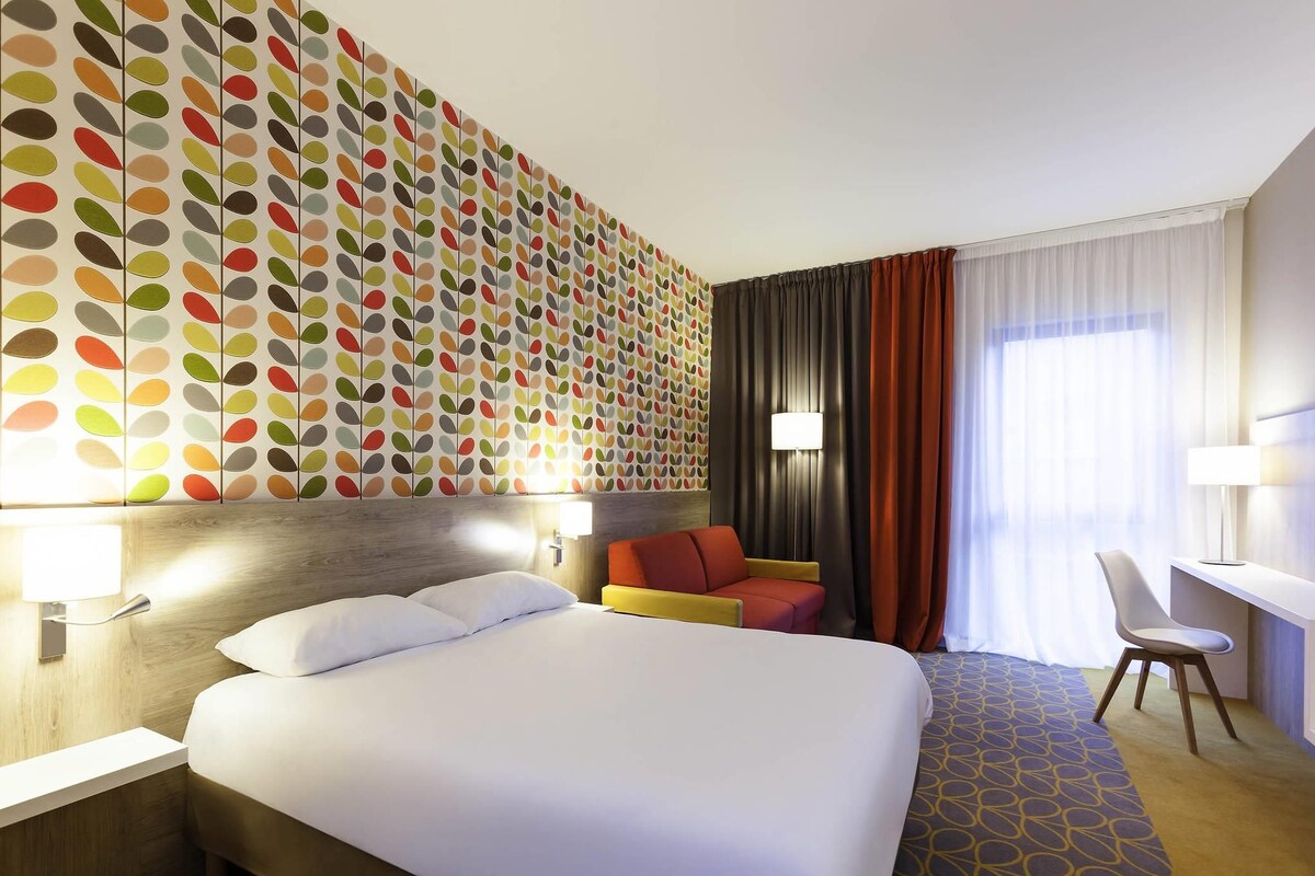 A hotel room with a large bed, a seating area with a chair, a desk, and a wall with a colorful leaf pattern.