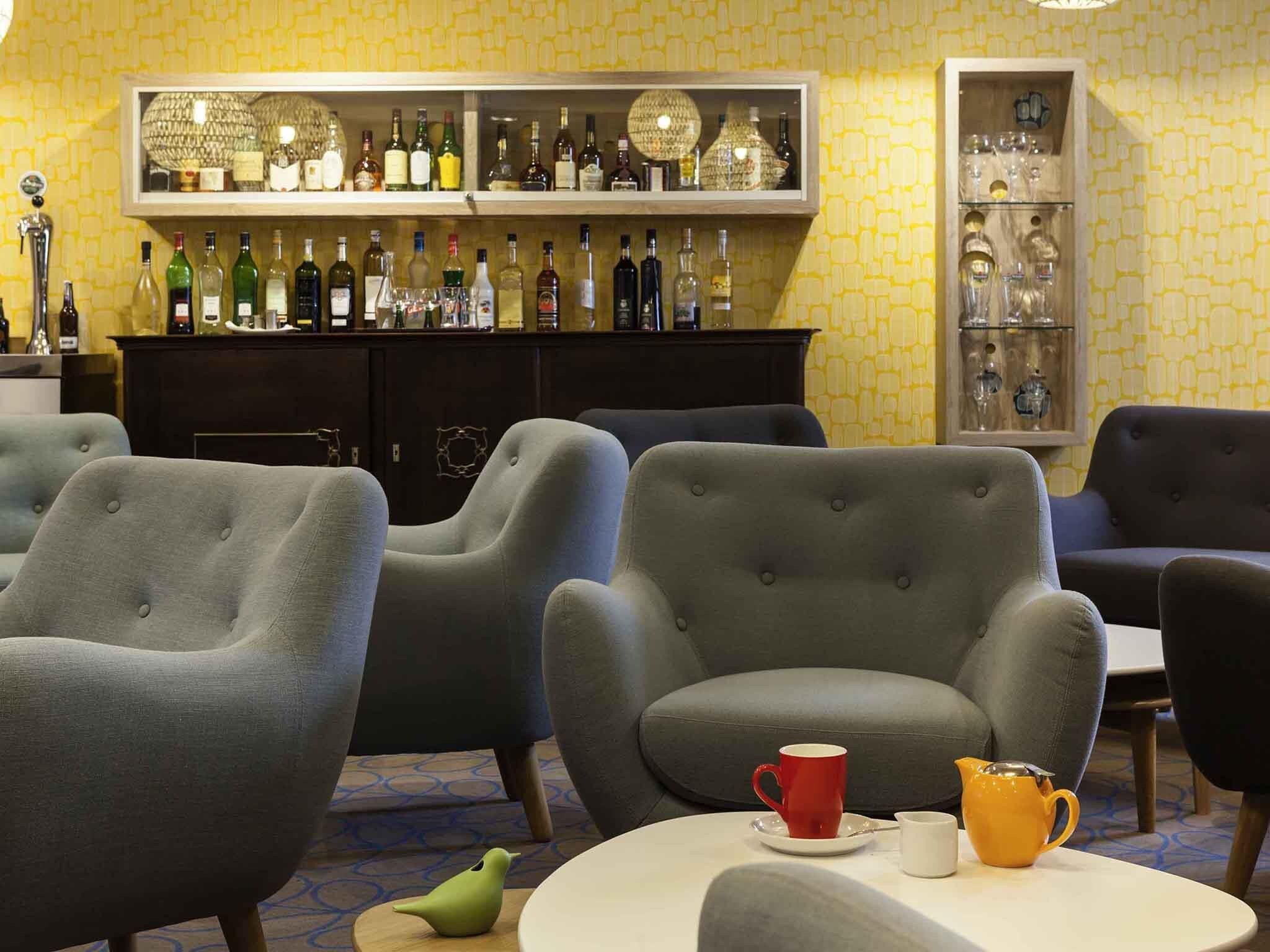A room with grey upholstered armchairs, a round table with a red cup and a yellow pitcher, and a bar area with various bottles and glasses.