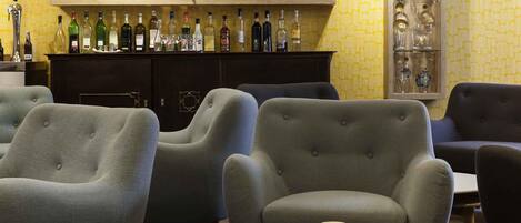 A room with grey upholstered armchairs, a round table with a red cup and a yellow pitcher, and a bar area with various bottles and glasses.