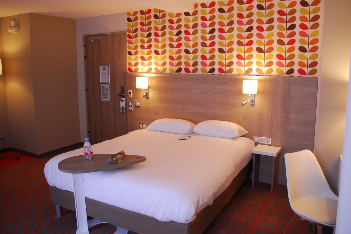 A hotel room with a bed, bedside tables, a chair, a wall with a leaf pattern, and a door.