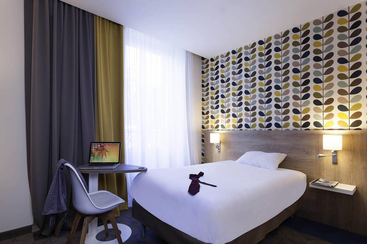 A hotel room with a bed, a desk with a laptop, a chair, and a wall with a leaf pattern.