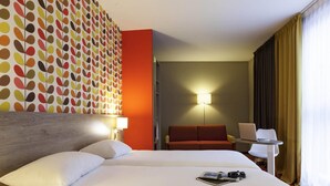 A hotel room with a large bed, a red sofa, a desk with a laptop, and a wall with a colorful leaf pattern.