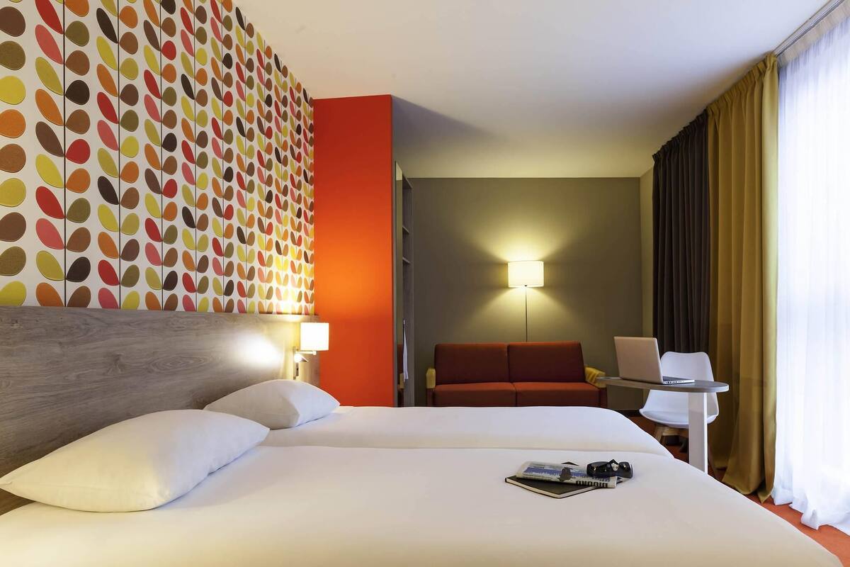A hotel room with a large bed, a red sofa, a desk with a laptop, and a wall with a colorful leaf pattern.