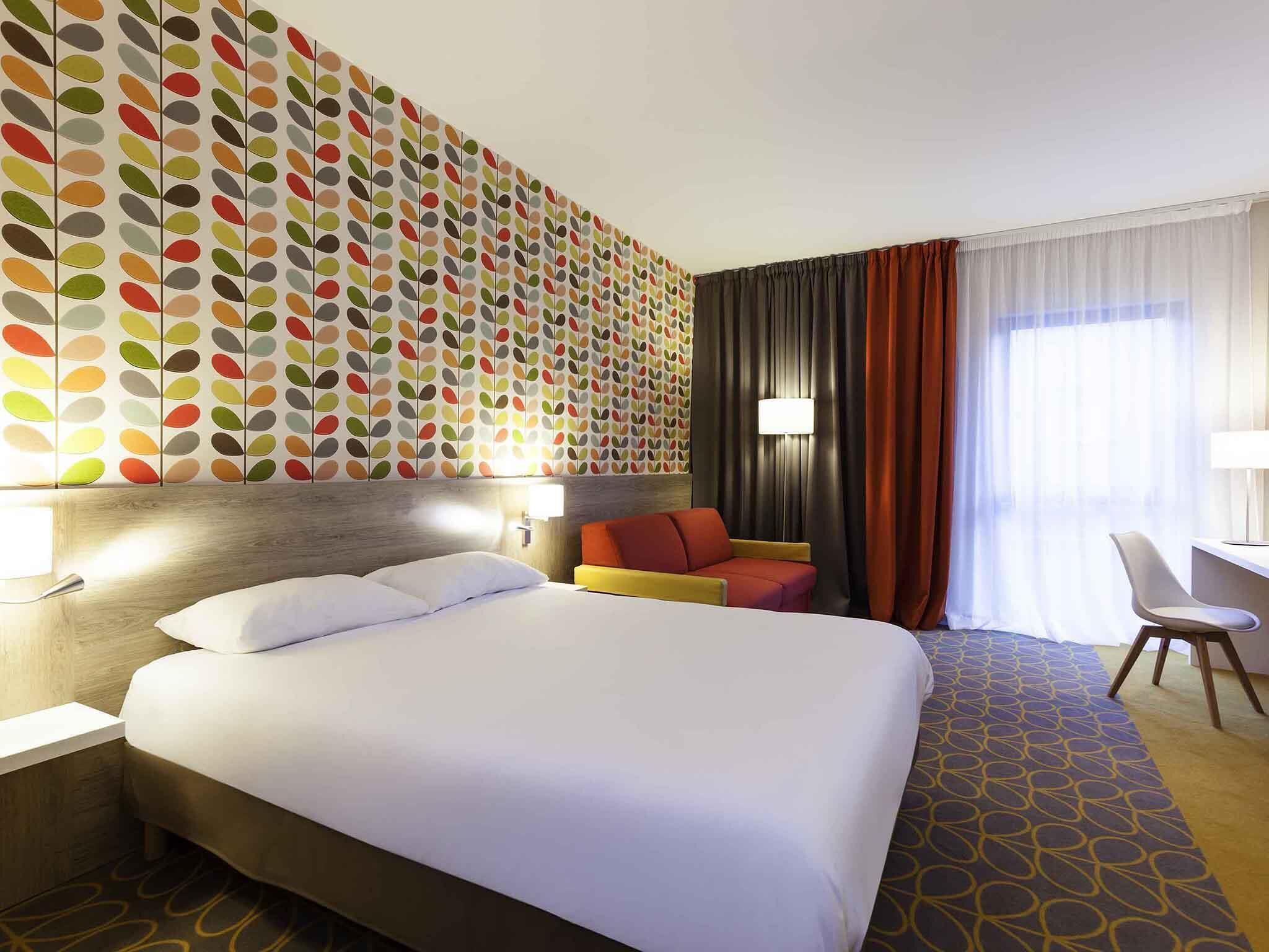 Habitación de hotel con una cama grande, un escritorio, una silla, una pared colorida y una ventana con cortinas.