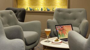A modern lounge area with grey upholstered chairs, a laptop on a small table, a glass of beer, and a decorative bird shelf in the background.