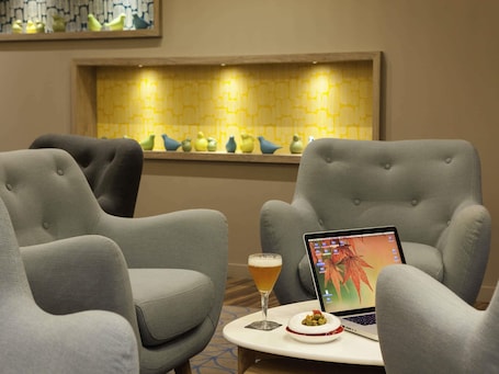 A modern lounge area with grey upholstered chairs, a laptop on a small table, a glass of beer, and a decorative bird shelf in the background.
