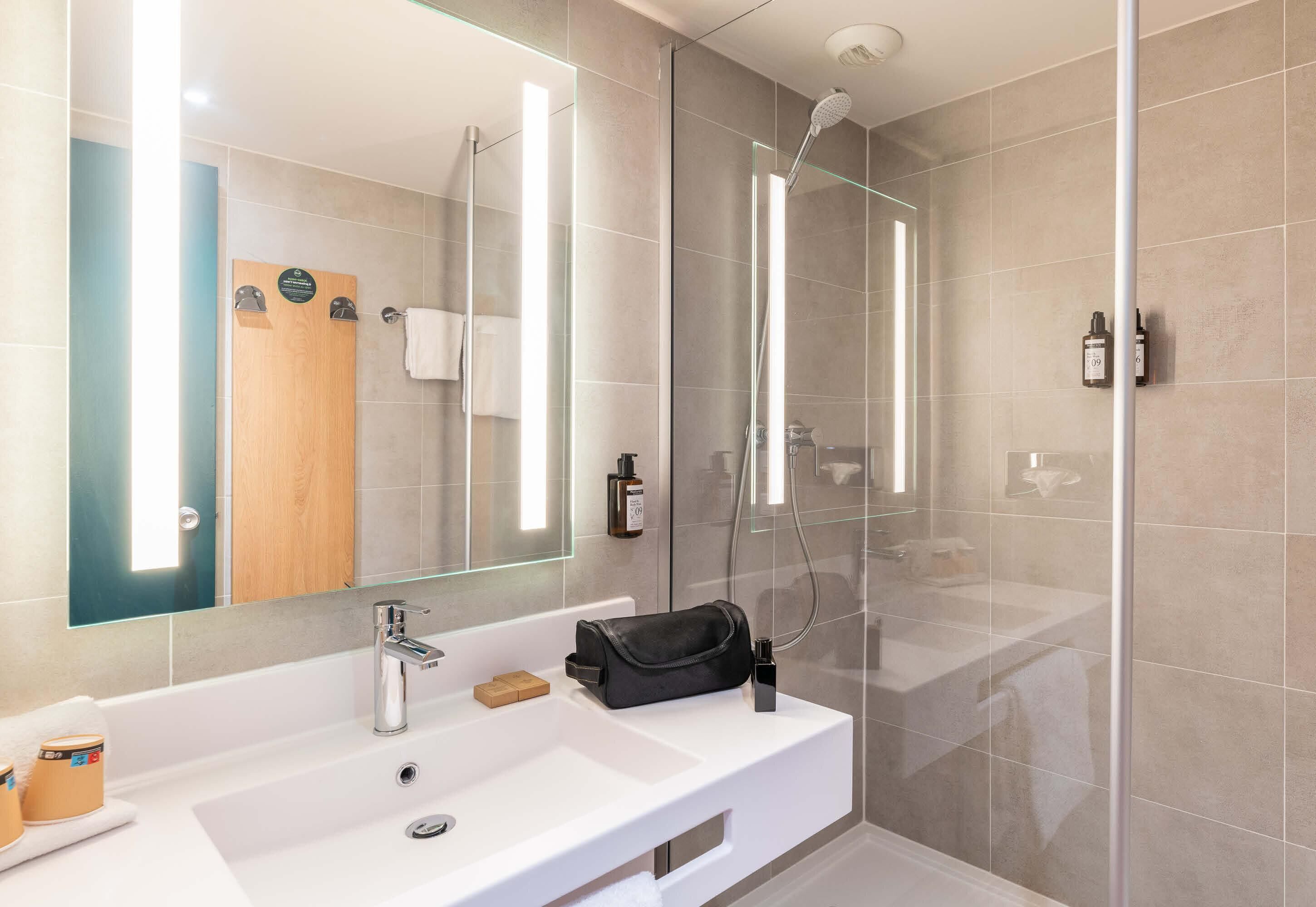 Une salle de bain moderne avec un grand miroir, un lavabo blanc, une douche à parois de verre et une porte en bois.