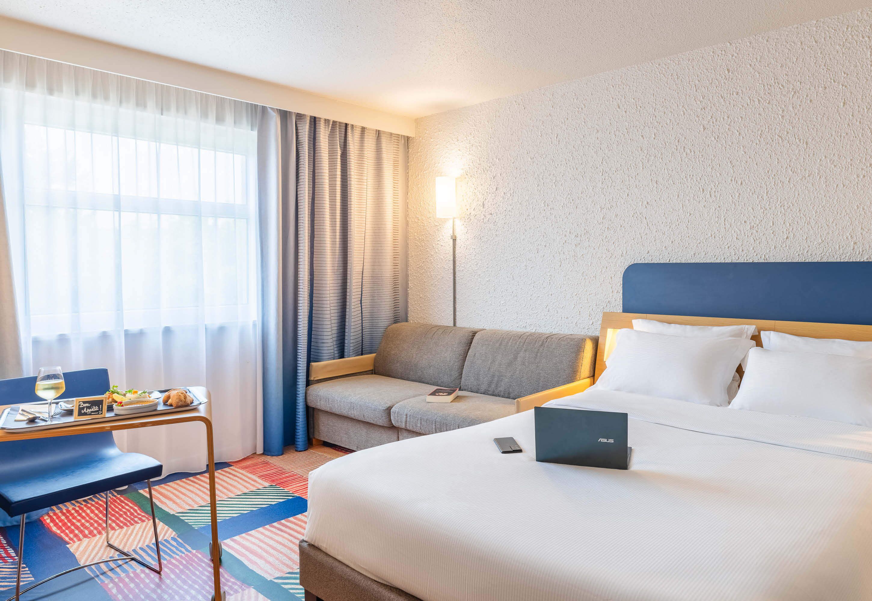 A hotel room with a bed, a desk with a laptop, a sofa, and a dining table with a glass of wine.