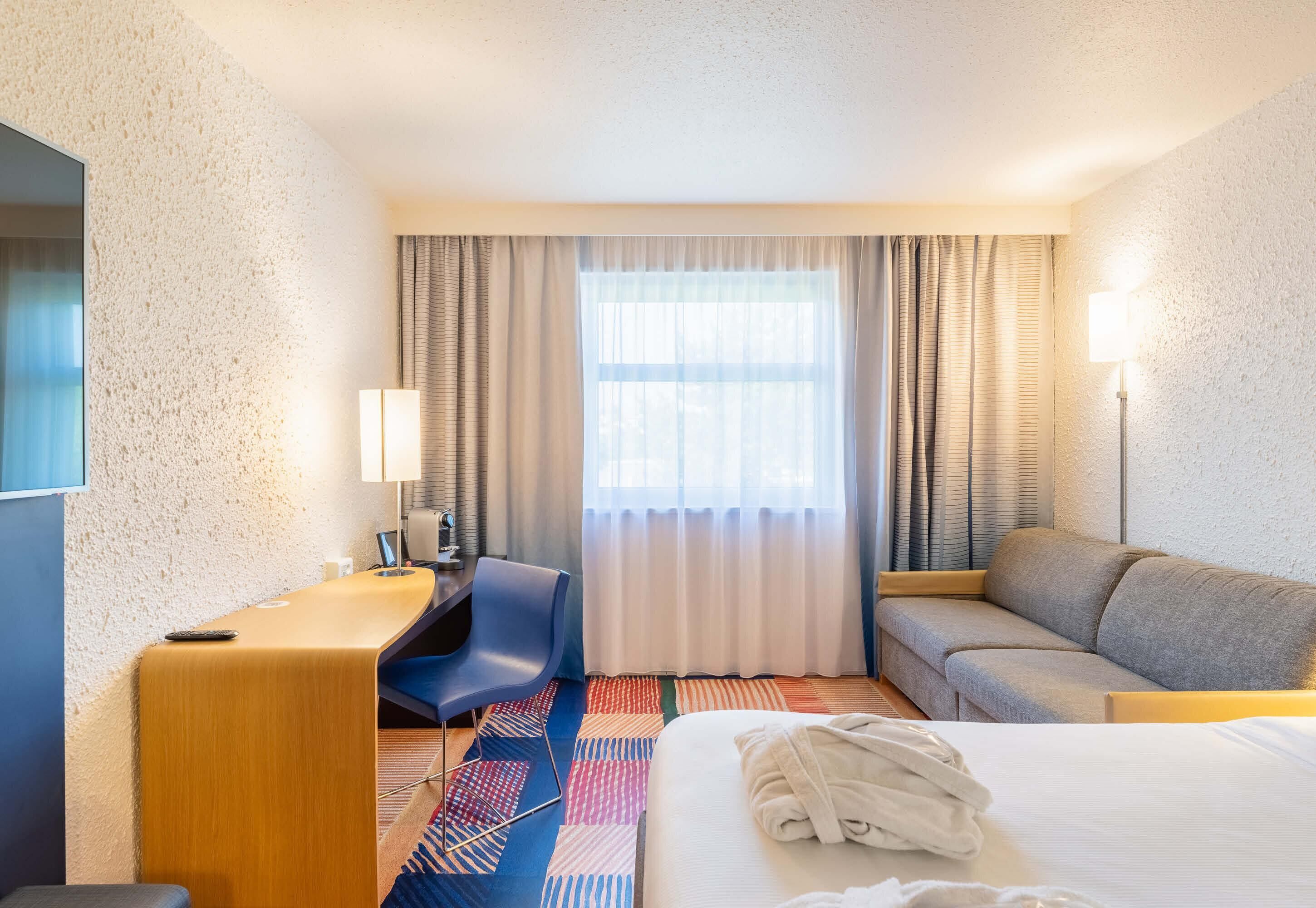 Quarto de hotel com cama, escrivaninha, cadeira, sofá e janela com cortinas.