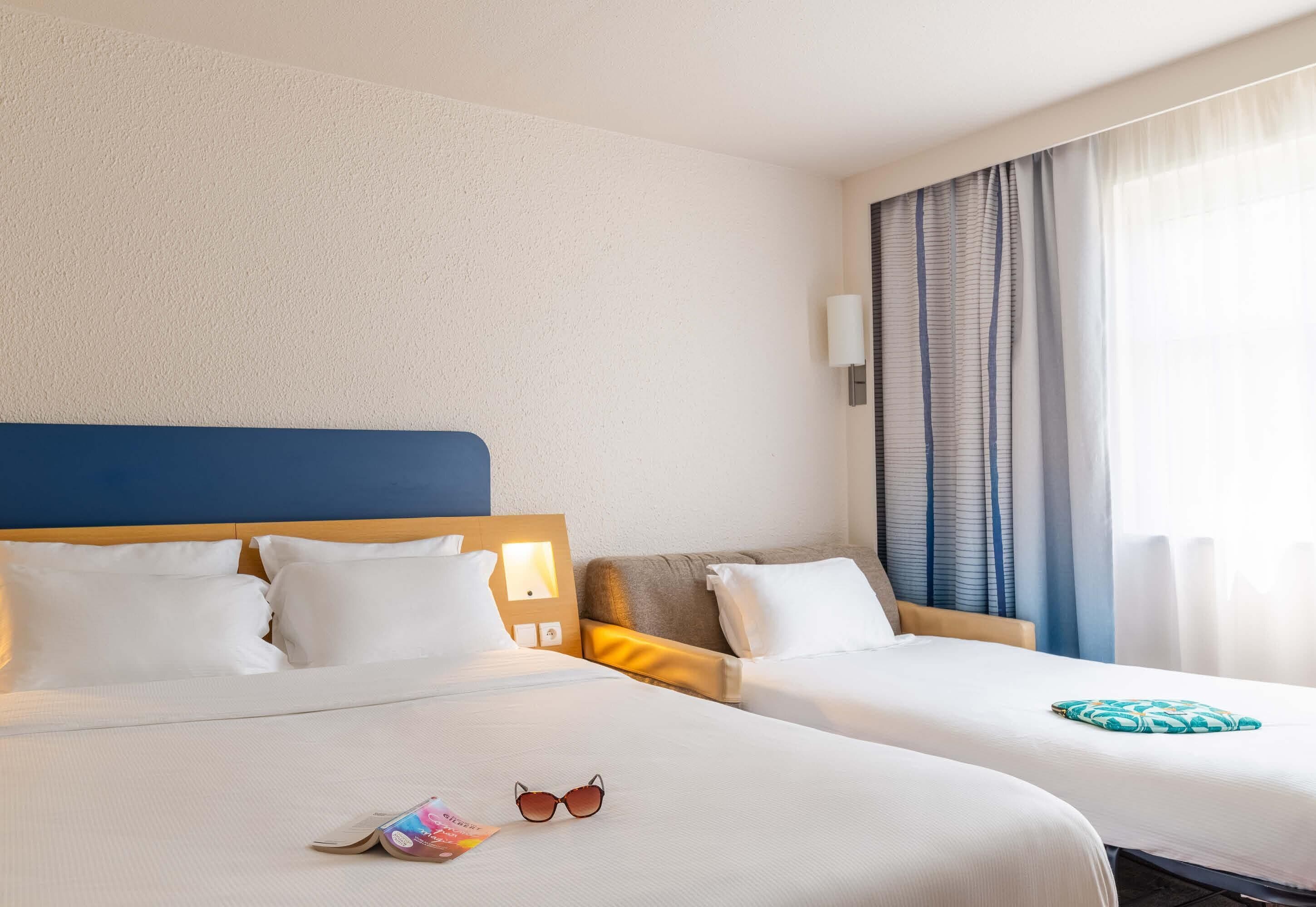 A hotel room with two beds, a headboard, a nightstand, a lamp, and a window with curtains.