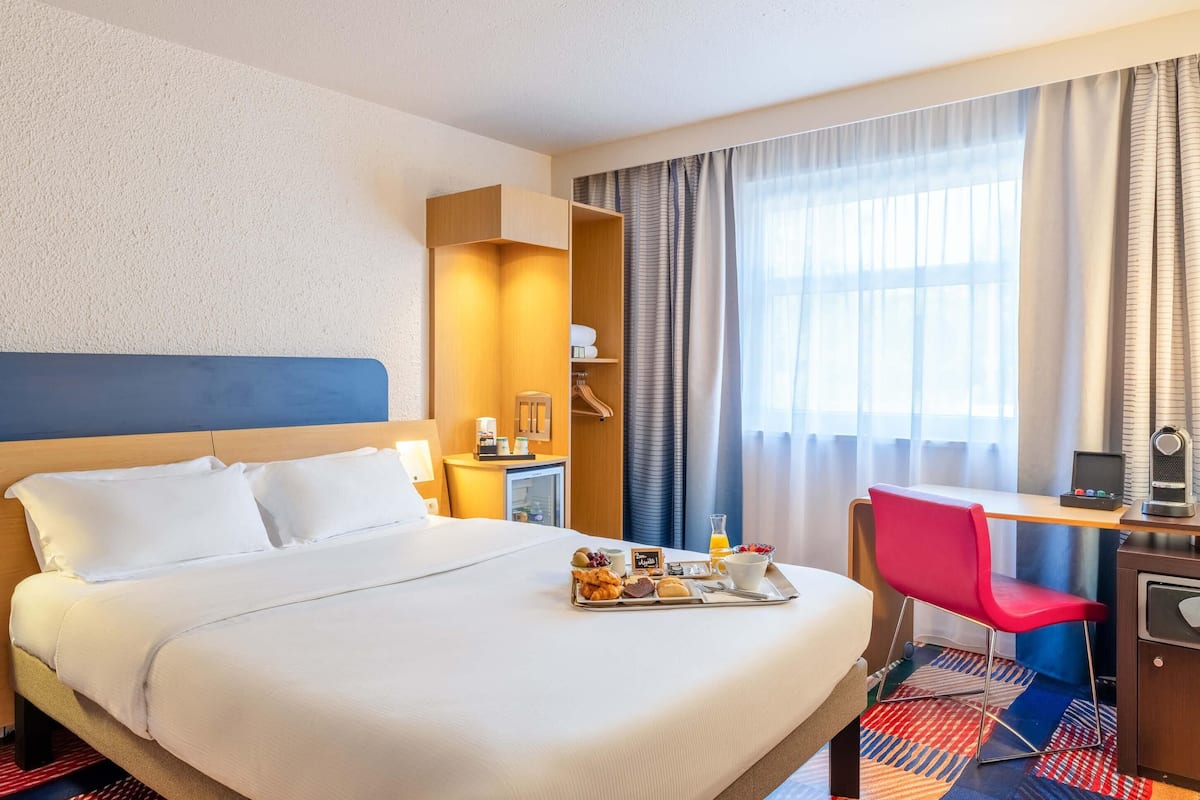 Ein Hotelzimmer mit Bett, Schreibtisch mit Stuhl, einer kleinen Küchenzeile und einem Fenster mit Vorhängen.