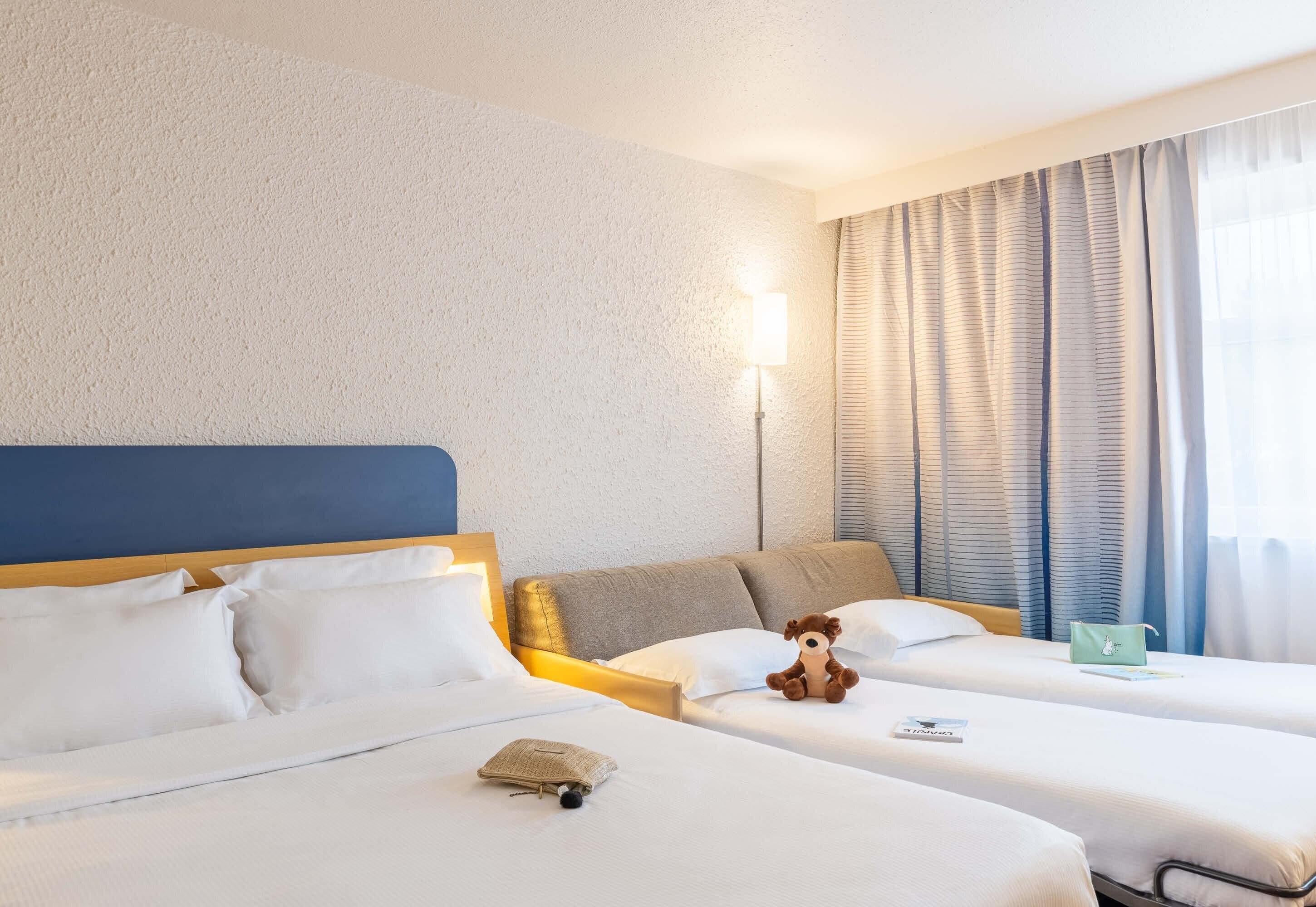 Une chambre d’hôtel avec deux lits, une tête de lit, une lampe, une fenêtre avec des rideaux et une petite table sur laquelle se trouvent un livre et un sac.