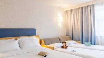 Habitación de hotel con dos camas, cabecero, lámpara, ventana con cortinas y una pequeña mesa con un libro y una bolsa.