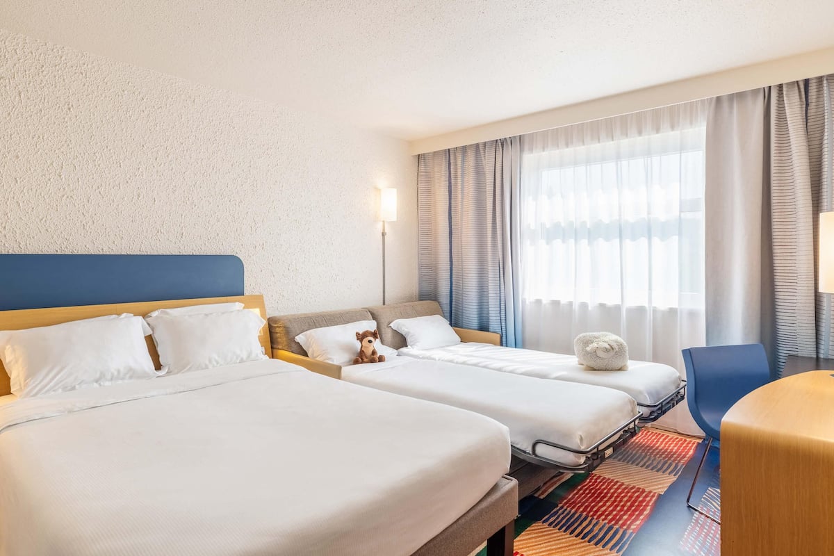 Ein Hotelzimmer mit zwei Betten, einem Schreibtisch, einem Stuhl und einem großen Fenster mit Vorhängen.