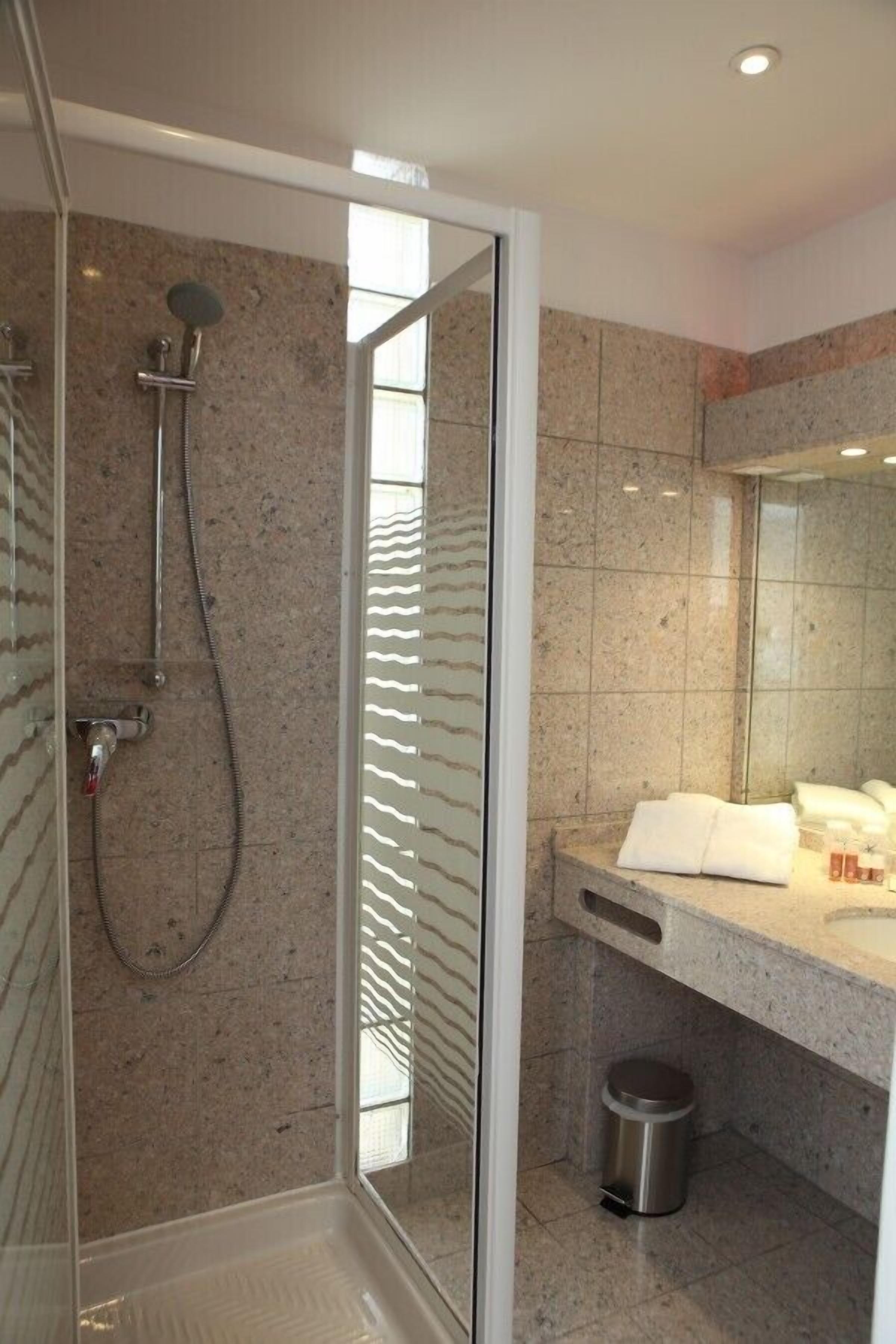 Une salle de bain moderne avec une douche à parois de verre, un mur recouvert de carreaux de marbre, un lavabo avec un miroir et une poubelle.