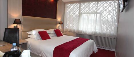 Habitación de hotel con una cama grande, dos mesitas de noche, un escritorio con una silla, un radiador y una ventana con celosía decorativa.