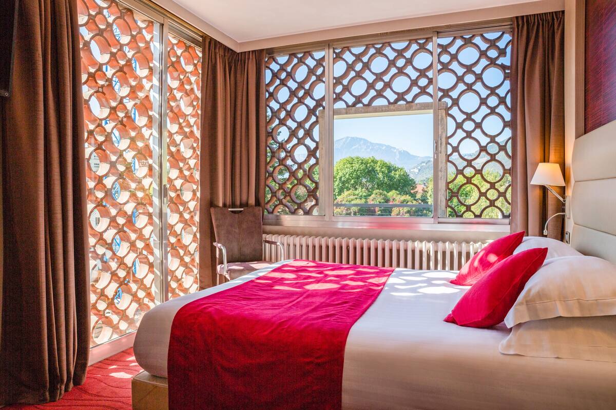A hotel room with a large bed, a chair, a window with a view of mountains, and a radiator.