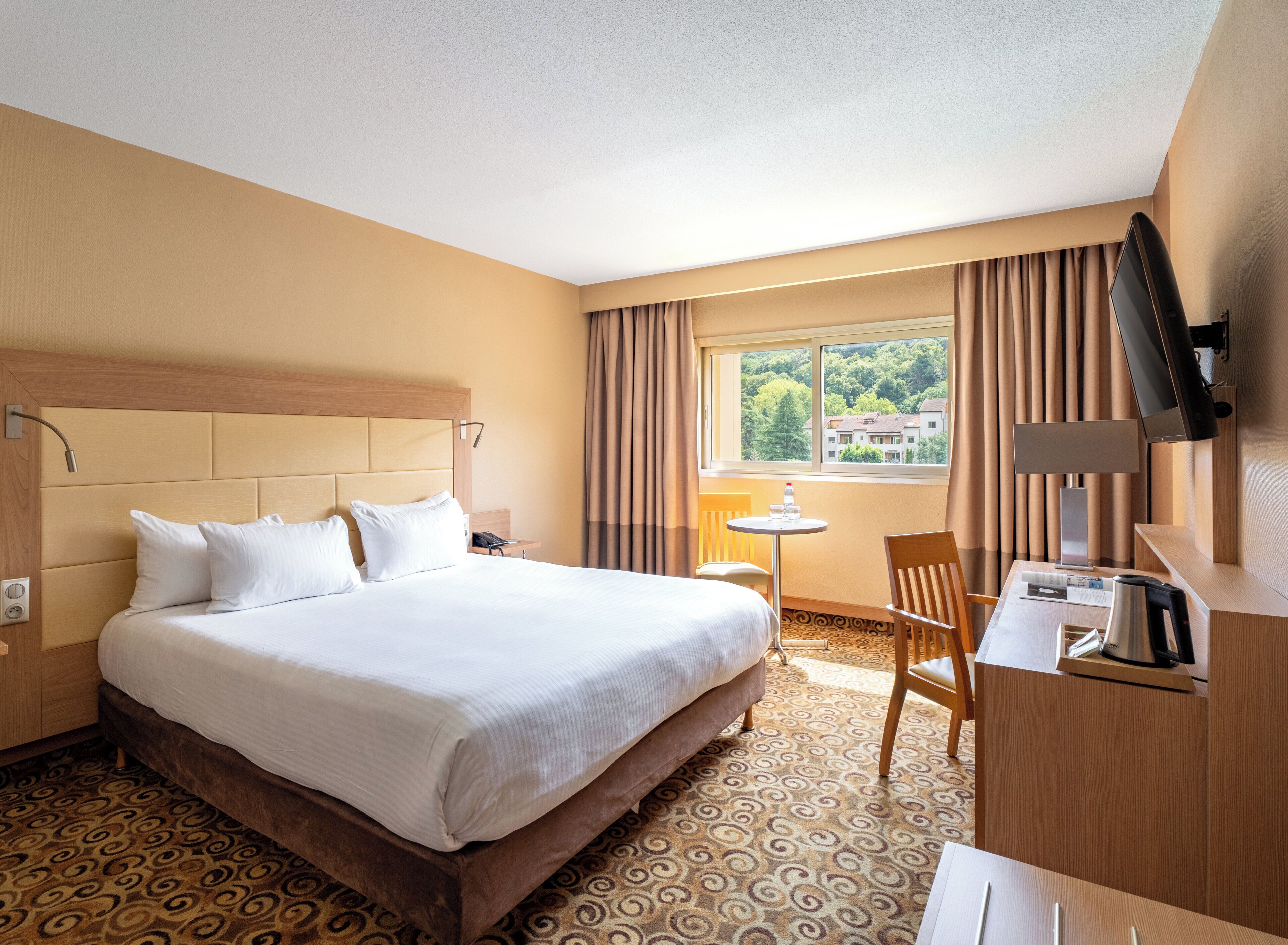 A hotel room with a bed, a desk, a chair, a TV, and a window with curtains.