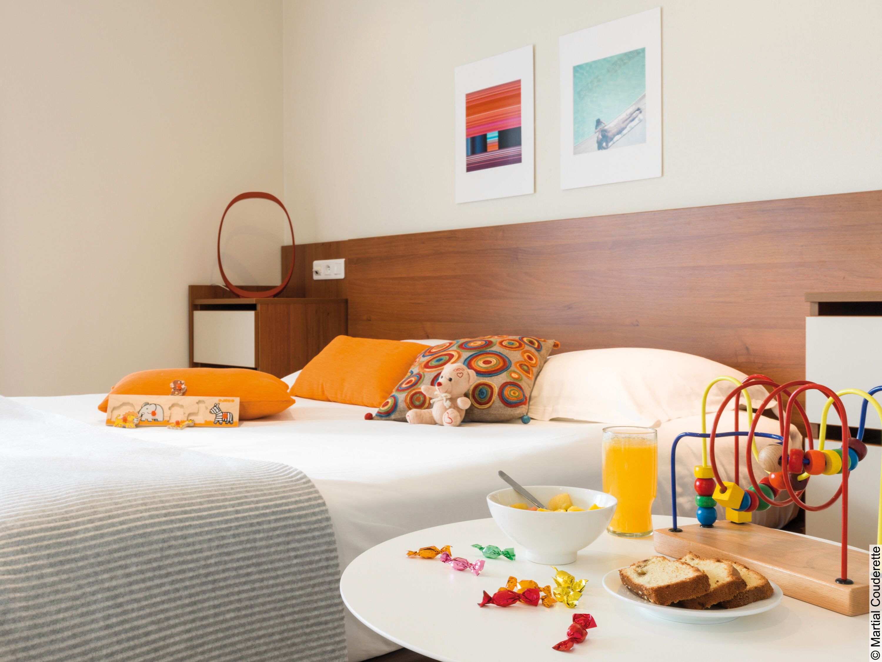 A hotel room with a bed, a bedside table with breakfast items, a wall with artwork, and a wooden headboard.