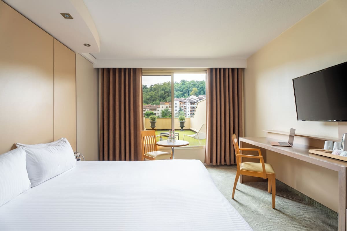 Une chambre d’hôtel avec un lit, un bureau avec un ordinateur portable, une télévision et une vue sur une cour intérieure agrémentée de plantes et de bâtiments.