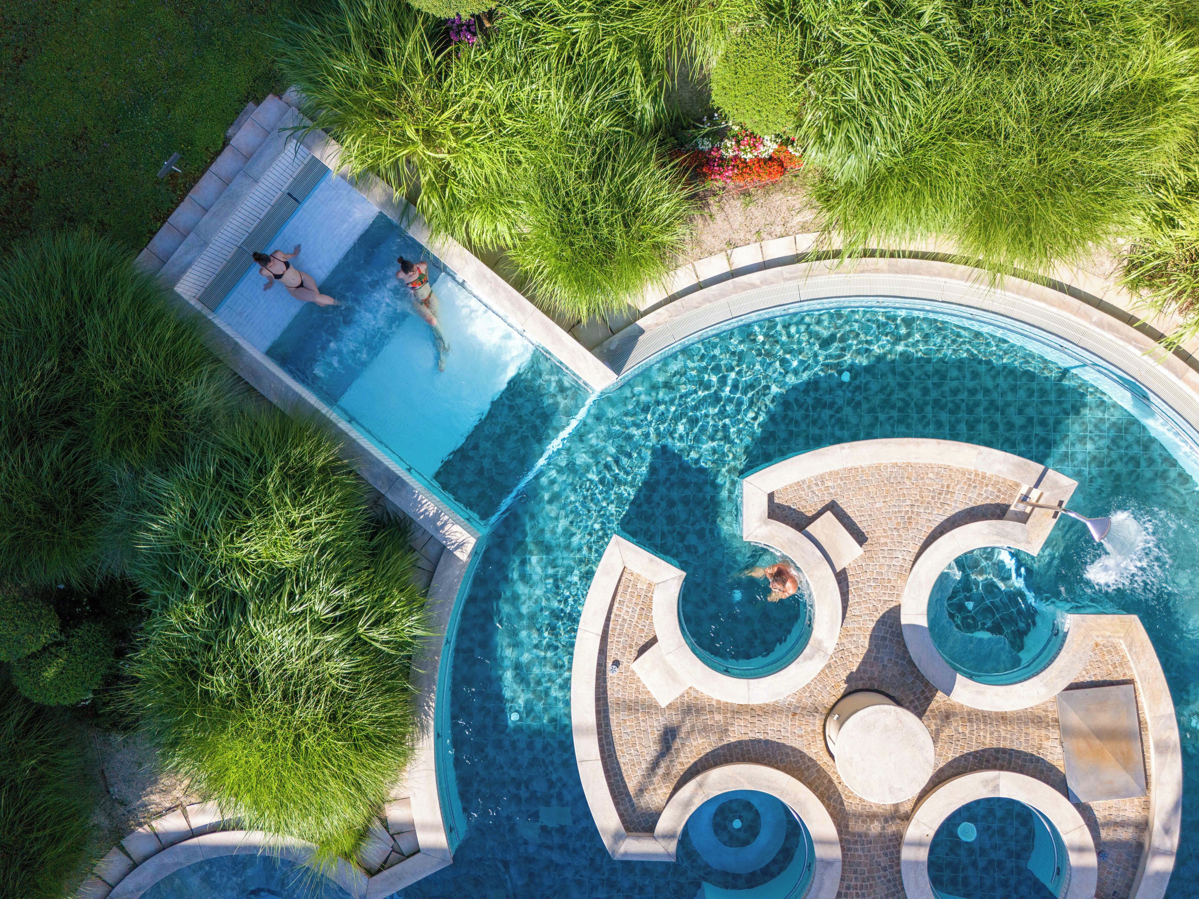 Una grande piscina a più livelli con acqua cristallina, circondata da verdeggiante vegetazione e un solarium piastrellato.