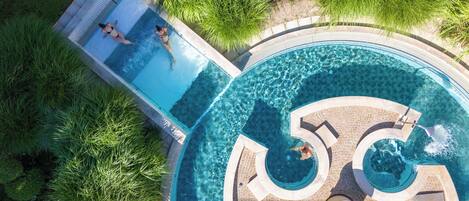Una grande piscina a più livelli con acqua cristallina, circondata da verdeggiante vegetazione e un solarium piastrellato.