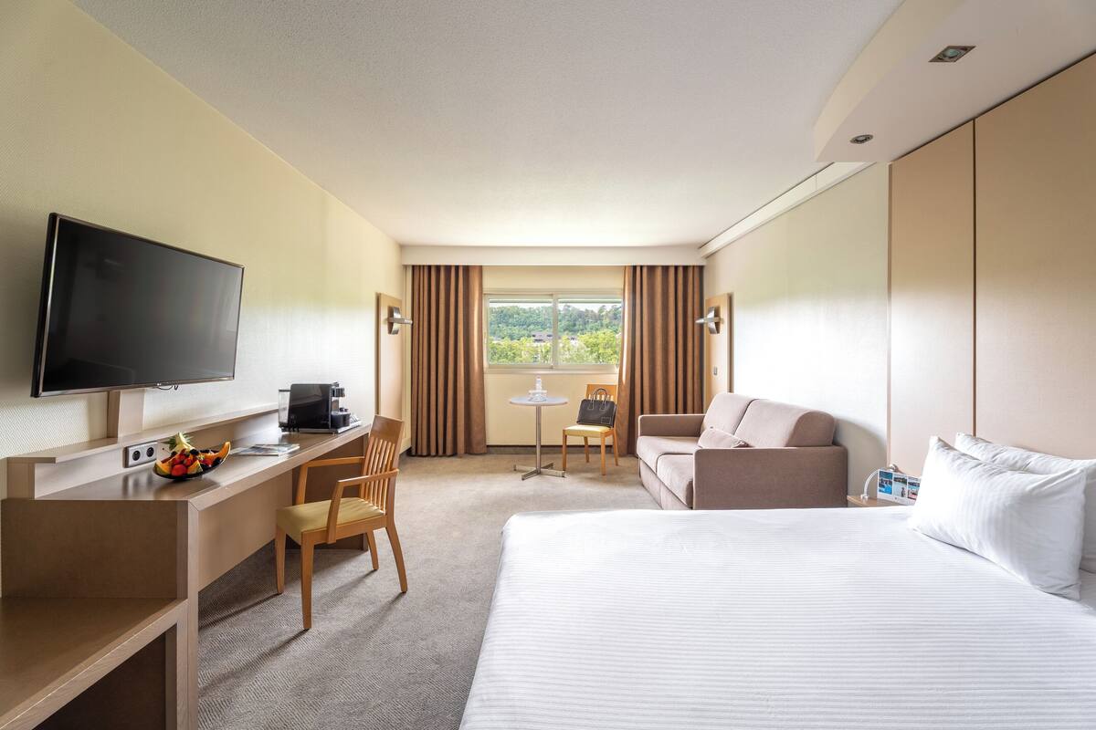 Une chambre d’hôtel avec un lit, un bureau avec une télévision, une chaise, une petite table et une fenêtre avec des rideaux.
