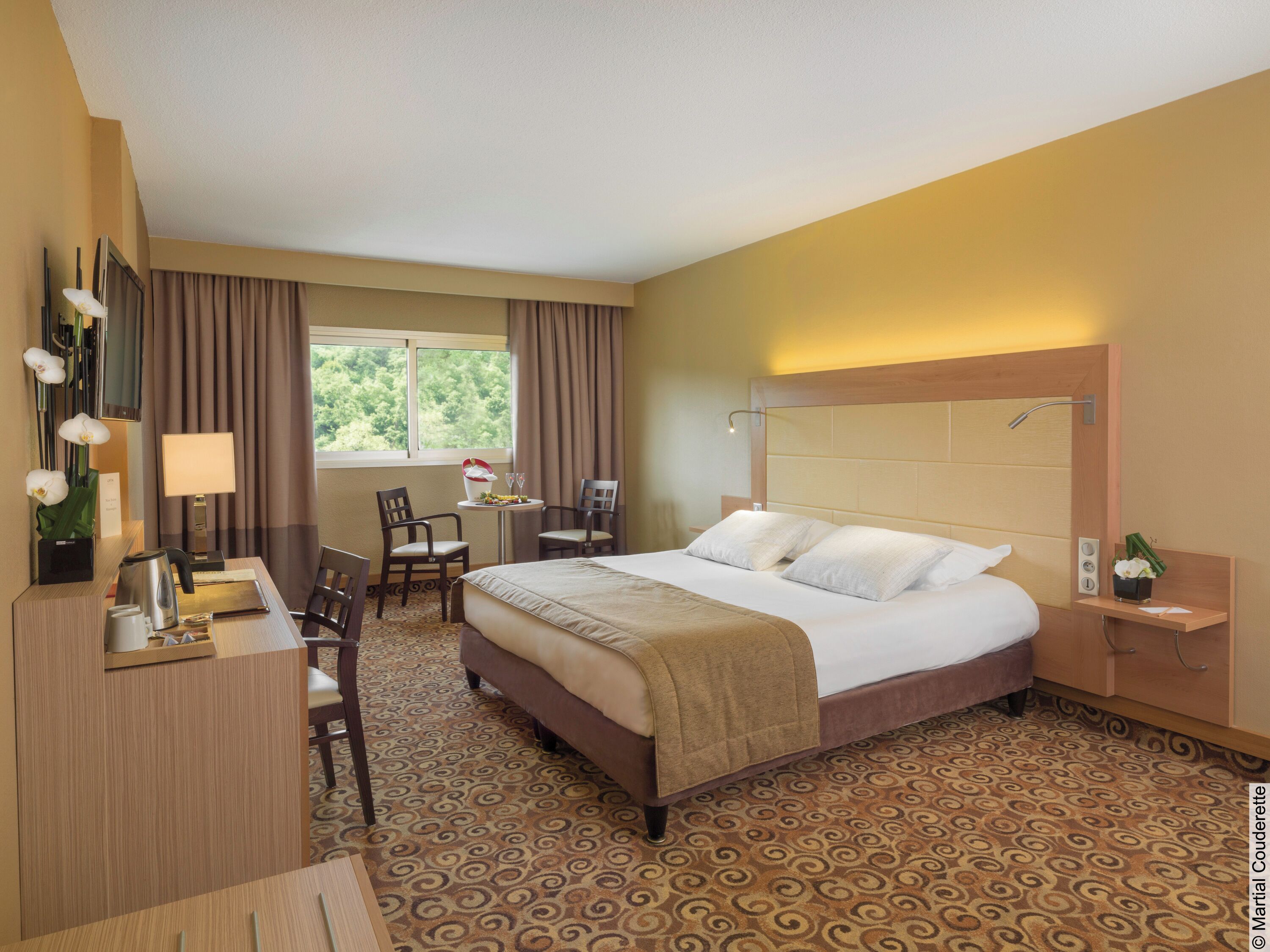 A hotel room with a bed, desk, chairs, a small table with a lamp, and a window with curtains.