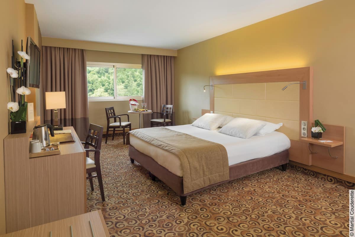 Une chambre d’hôtel avec un lit, un bureau, des chaises, une petite table avec une lampe et une fenêtre avec des rideaux.