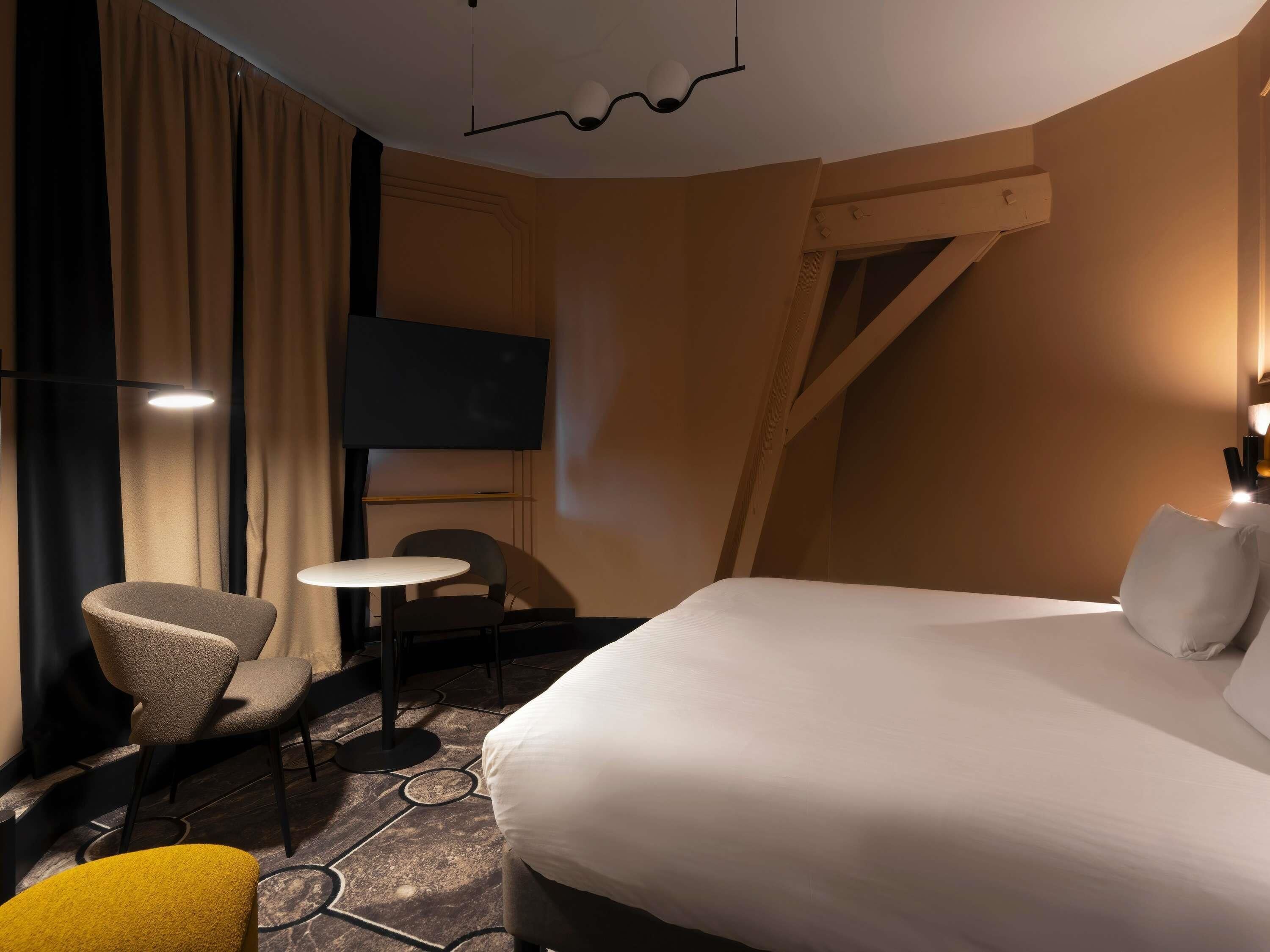 A hotel room with a bed, a TV, a small table, two chairs, and a yellow ottoman.