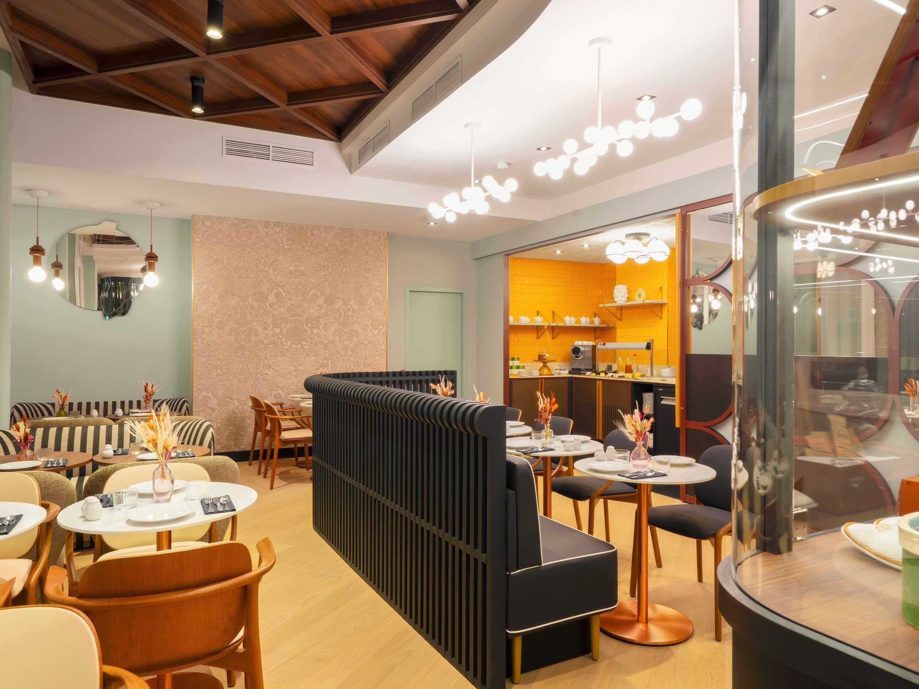 A modern restaurant with a mix of seating options including booths and tables. There is a visible kitchen area with a counter and various kitchen equipment. The ceiling features exposed wooden beams and recessed lighting.