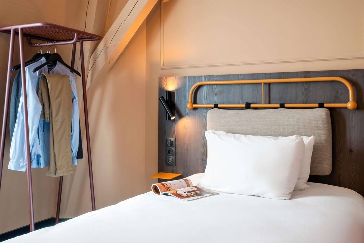 Una habitación de hotel con una cama, cabecero de madera, mesita de noche, lámpara y un perchero con ropa colgada.