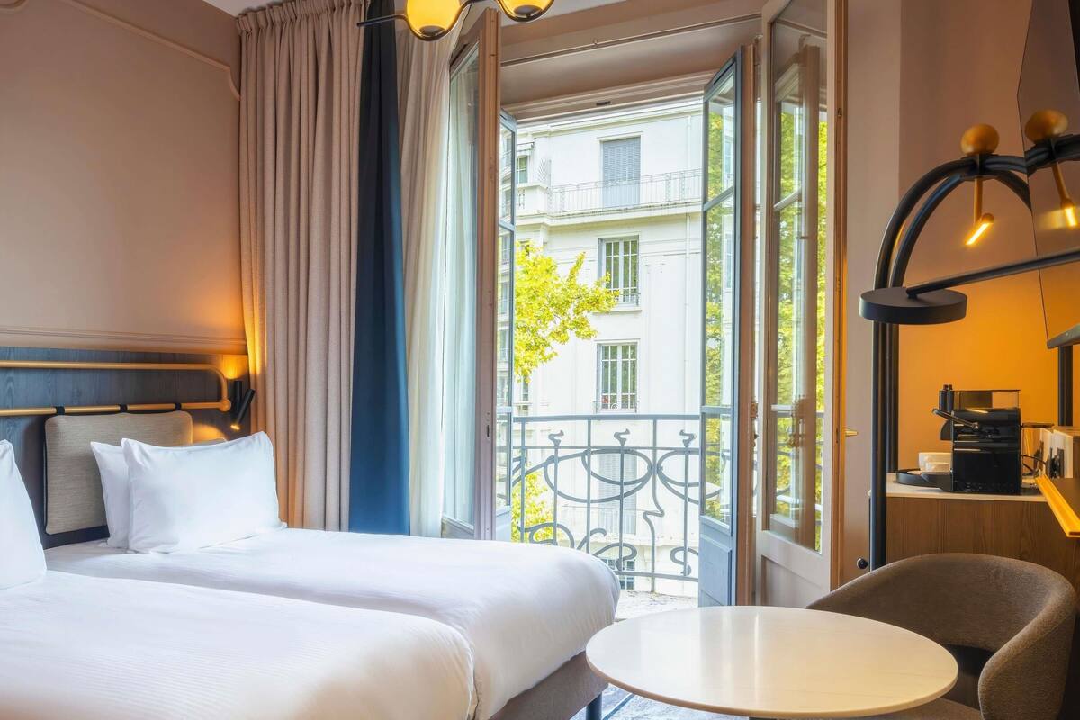 Habitación de hotel con dos camas, una mesita redonda pequeña, una silla, un balcón con vistas a edificios y una cafetera.