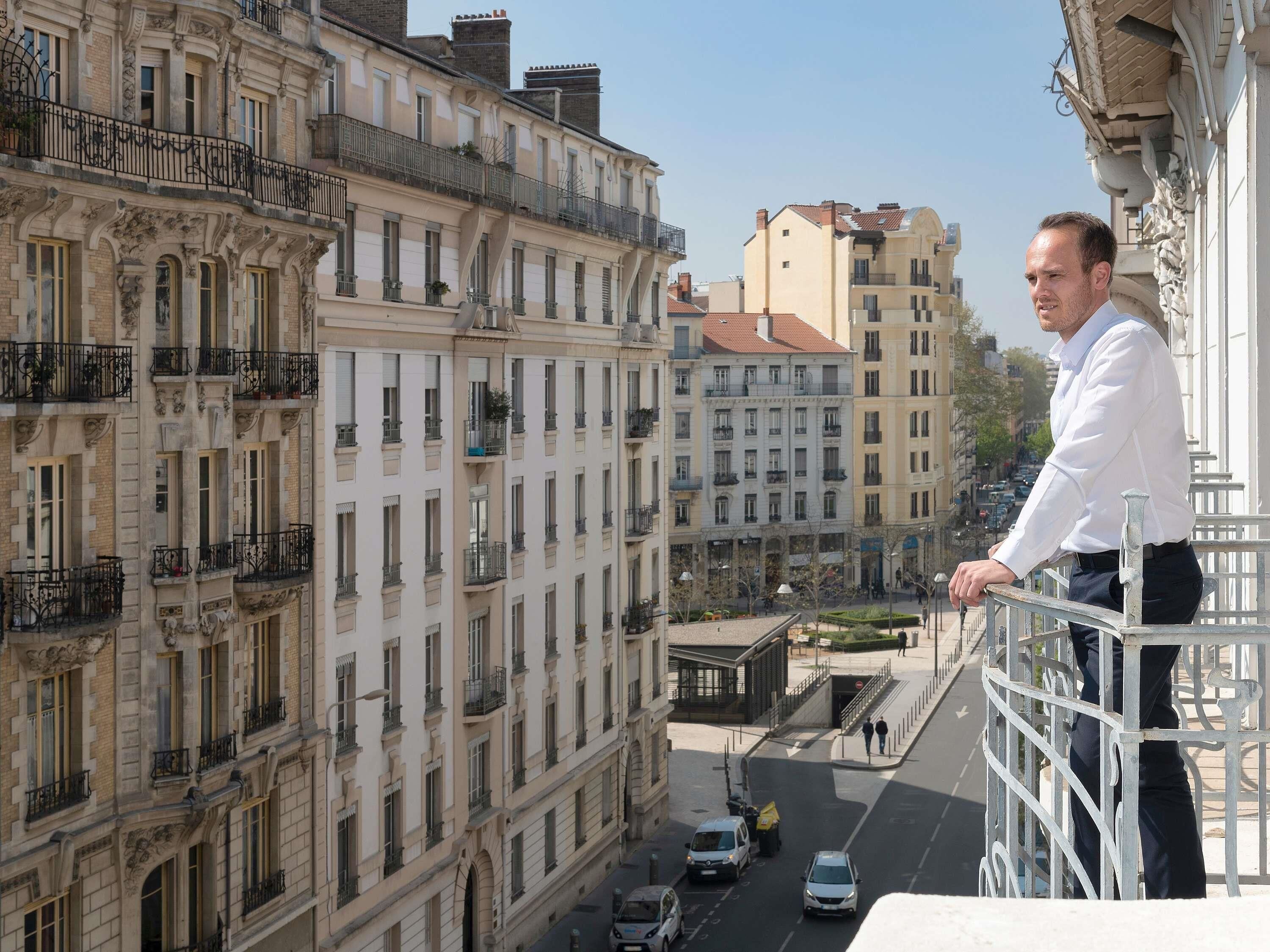 A man standing on a balcony overlooking a city street with buildings and parked cars.