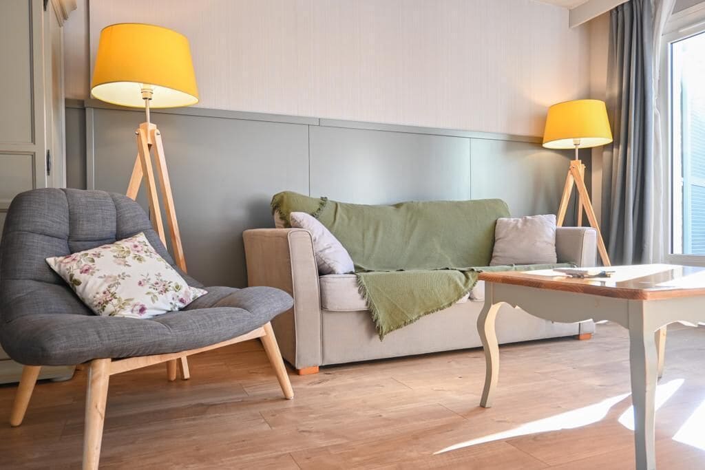 A modern living room with a grey armchair, a grey sofa, and a wooden coffee table. There are two floor lamps with yellow shades.