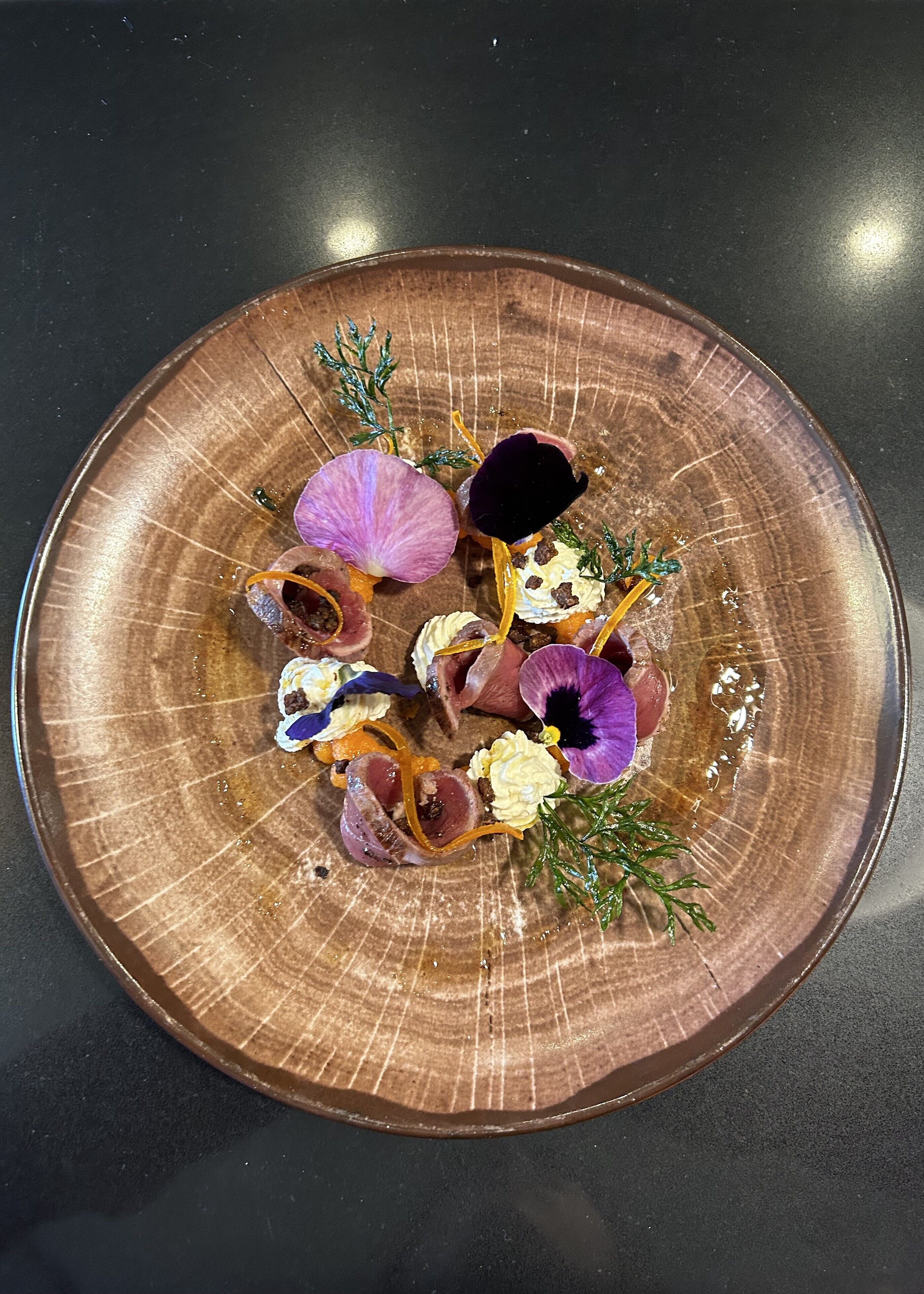 A round, textured brown plate with various food items and garnishes, including purple flowers and green herbs.